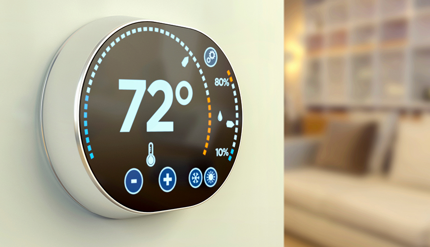A modern, circular digital thermostat displaying 72 degrees Fahrenheit. The screen shows temperature controls and a partial humidity bar. The thermostat is mounted on a wall in a room with dim lighting and a blurred background.