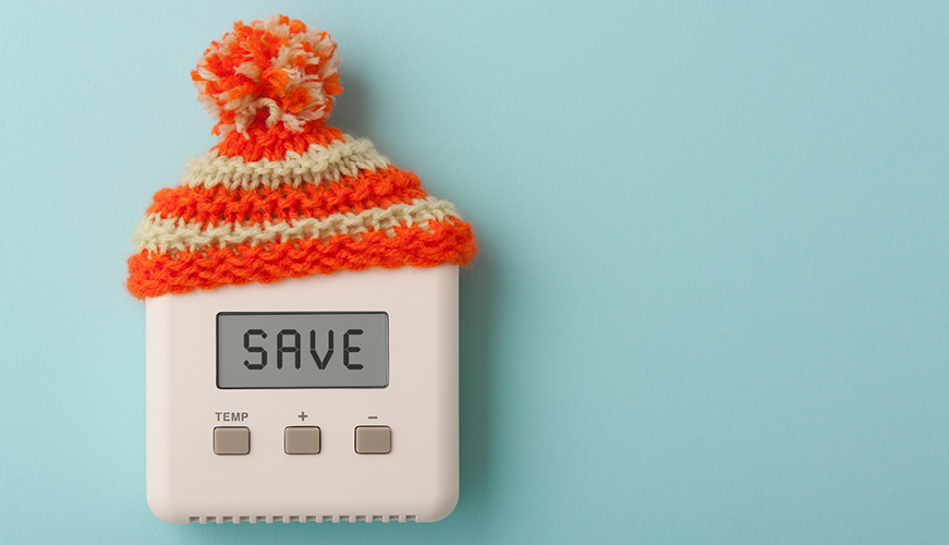 A thermostat with an orange and white knit hat is displayed against a light blue background. The digital screen reads "SAVE," emphasizing energy conservation.