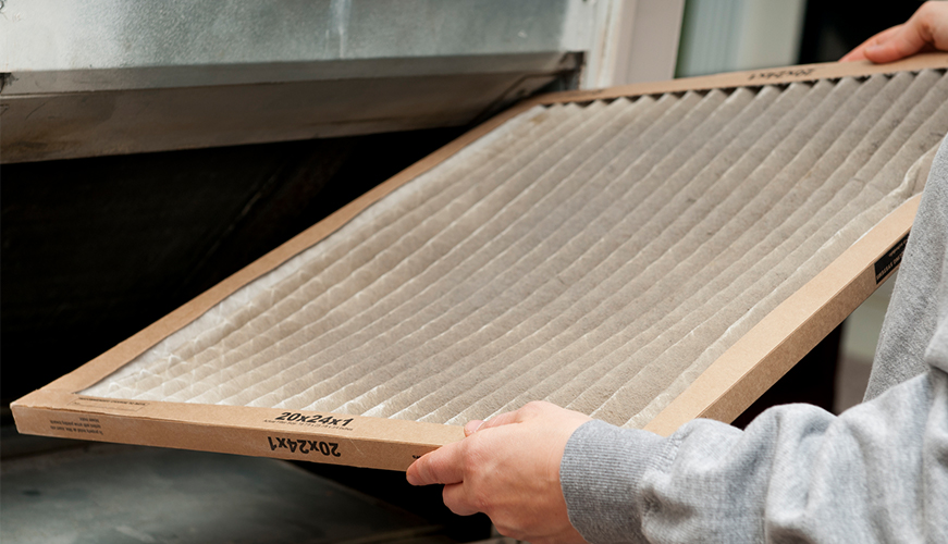 A person holds a dirty heating, ventilation, and air conditioning (HVAC) air filter and is preparing to replace it. The filter is rectangular with a cardboard frame and appears dusty, indicating it needs maintenance. It is held in front of a vent.