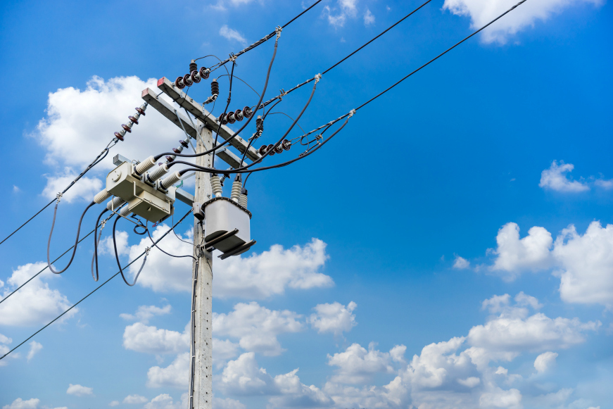 A utility pole with transformers and multiple electrical wires stands against a bright blue sky filled with scattered white clouds.